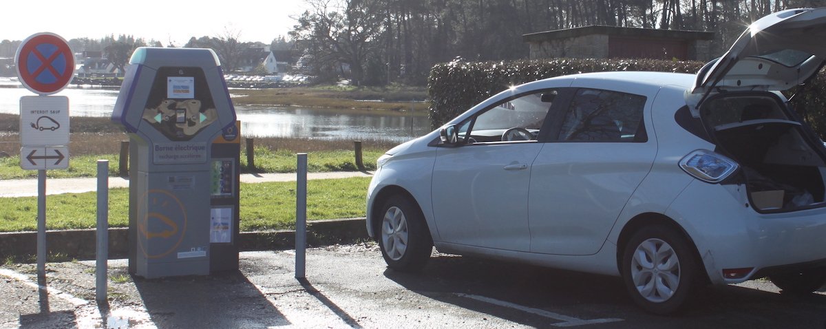 Photographie d&#x27;une borne de recharge électrique prise à Vannes à Conleau. Une voiture électrique blanche est sur la place prête à être rechargée.